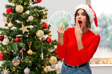 winter holidays and finance concept - surprised young woman in santa helper hat holding euro money...