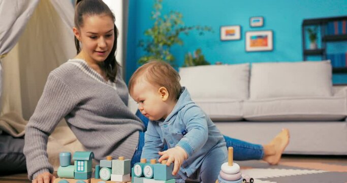 A smiling woman is playing on mat in the living room with her little son, boy is crawling, pushing a train made of wooden elements, is interested in toys, takes them to his mouth
