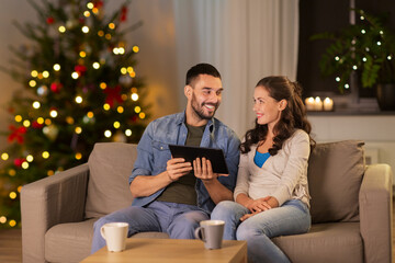 Obraz premium technology, winter holidays and people concept - happy couple using tablet computer at home in evening over christmas tree lights on background