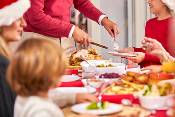 Grandfather Carving Turkey As Multi-Generation Family Sit Down To Eat Christmas Meal Together