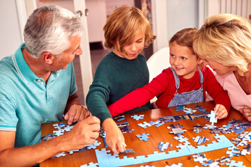 Grandchildren With Grandparents Sit Around Table At Home Doing Jigsaw Puzzle Together