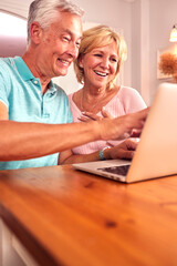 Senior Retired Couple Sitting On Sofa At Home Shopping Or Booking Holiday On Laptop