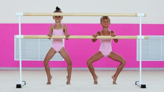 Two little girls gymnasts warming up in the studio - squatting standing on their tiptoes and holding by a handrail