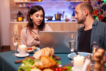 Cheerful couple buying xmas gift present paying online with credit card on laptop computer sitting at dining table in x-mas decorated kitchen. Happy family shopping for winter holidays