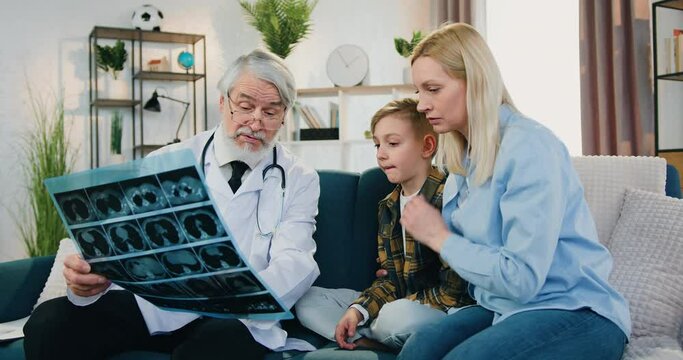 Accelerating shooting of attractive satisfied adult mother which discussing with experienced mature bearded doctor results of x-ray scan of her teen son during doctor's home visit