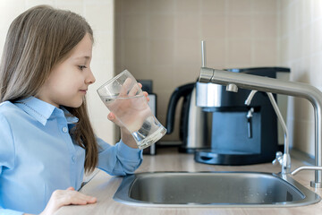 A glass of clean water in kids hands. Caucasian little girl drinking from water tap or faucet in...