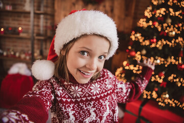 Photo of adorable blogger little kid girl make selfie showing evergreen tree wear ornament pullover santa hat indoors