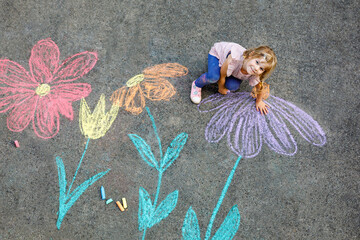 Little preschool girl painting with colorful chalks flowers on ground on backyard. Positive happy...