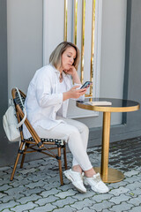 Young woman in a cafe works on a smartphone while sitting in a cafe. Blond business woman in white clothes.