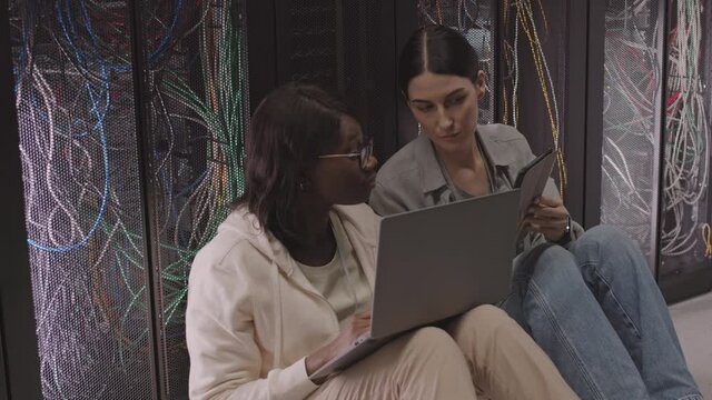 High angle of two multiethnic female IT specialists leaning back on server racks sitting on floor, using laptop and tablet computers, talking