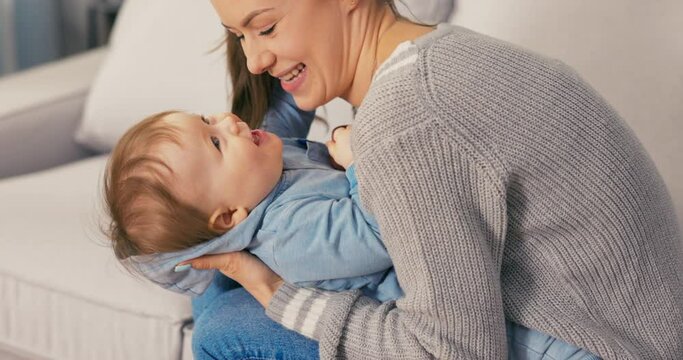 An attractive adult mom sits on living room couch with child in arms, which she lowers down grasping tightly and tickling, kissing boy's neck, playing with him, teasing, son laughing, happy