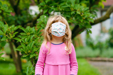 Little toddler girl on her first day on way to playschool with medical mask against corona covid virus. Preschool child walking to nursery preschool and kindergarten, making fun with mask, hiding face