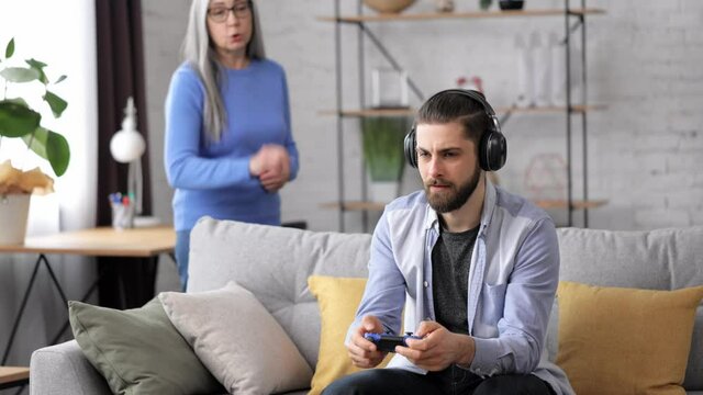 Two generation family. Unemployed adult son playing video games at home using gamepads ignoring senior mother yelling at him. Video game addiction. Family and generations conflicts, misunderstanding.
