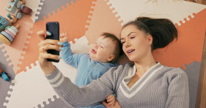 Young attractive mom lies on floor with son dressed in a blue bodysuit, boy looks around with big beautiful eyes, woman records video, takes picture for social media, brags about her child