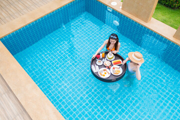 Top view of two women eating breakfast on floating tray in swimming pool in the luxury villa...