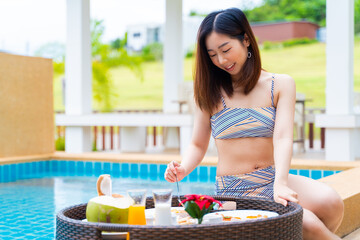 Portrait of Asian woman in swimwear enjoys floating breakfast tray in the luxury hotel resort. Female relaxing in summer vacation.