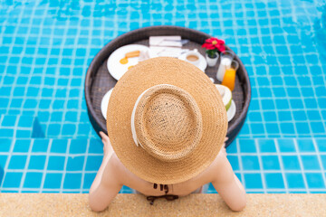 Rearview of woman in sun hat relaxing in swimming pool with floating breakfast tray at luxury hotel. Female enjoying summer vacation outdoors.