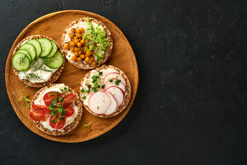 Four crispy buckwheat bread with cream cheese, radish, tomato, chickpea, cucumber and microgreen for healthy breakfast on parchment paper on black stone background. Concept vegan and healthy eating.
