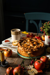 Autumn still life with fruit pie, decorated with crust apple pattern, apples,  lingonberries, candle and cup of apple tea on old wooden table and chair on background. 