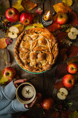 Autumn fruit pie decorated with crust apple pattern, apples,  lingonberries, candle, fallen leaves and woman hands holding cup of apple tea on old wooden table. 