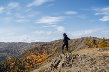 A girl in dark clothes walks along the top of the mountain, looks at the mountains and forest, the clear sky overhead, the autumn landscape.