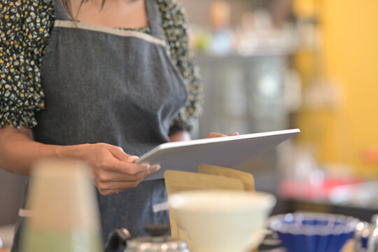 Cropped image of a young woman wearing an apron is holding a digital tablet while standing behind the bar counter in a coffee shop. Young business owner concept.