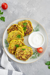 Vegetable zucchini pancakes with cheese, dill and parsley on a plate with sour cream on a gray concrete background. Top view, copy space.
