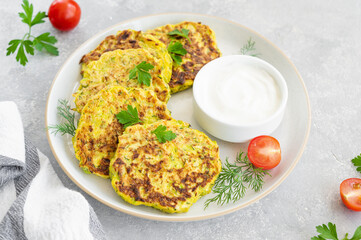 Vegetable zucchini pancakes with cheese, dill and parsley on a plate with sour cream on a gray concrete background. Top view, copy space.