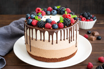 Three chocolate mousse cake with chocolate glaze, blueberries, raspberries and blackberries on top on a white plate on a dark wooden background. Cake with three layers of different taste. Copy space.