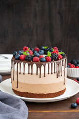 Three chocolate mousse cake with chocolate glaze, blueberries, raspberries and blackberries on top on a white plate on a dark wooden background. Cake with three layers of different taste. Copy space.
