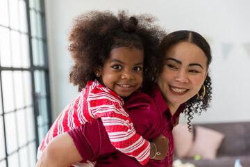Happy African American mother piggybacking with cute curly little daughter on back. Cheerful...