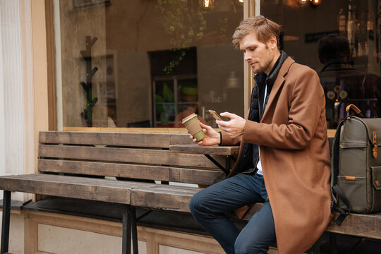 Handsome man holding takeaway coffee cup and mobile phone in hand. City life cafe in background. Man in brown coat Modern businessman with backpack