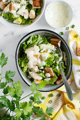 Traditional caesar salad in a white bowl on a old white table with leaves and a sauce boat and parmesan. Selected focus.