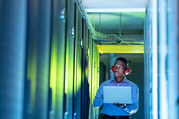 African american male computer technician wearing headphones using laptop working in server room