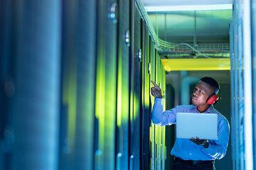 African american male computer technician wearing headphones using laptop working in server room