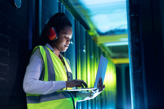 African american female computer technician wearing headphones using laptop working in server room