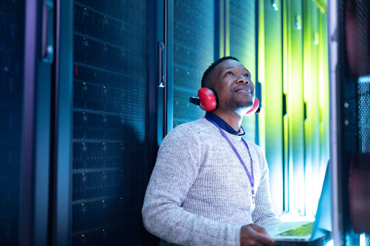 African american male computer technician wearing headphones using laptop working in server room