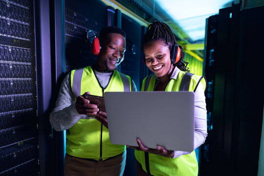 Happy african american computer technicians wearing headphones using laptop working in server room