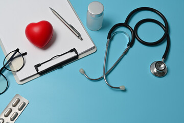 Top view of doctor desk with a stethoscope, capsule pills, bottle of medicine, glasses, heart model, pen and blank paper on the clipboard. Top view with copy space, flat lay.