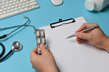 Top view of the doctor's hands is writing on a clipboard over a blue background surrounded by a stethoscope, wireless keyboard, mouse, and bottle of medicine.