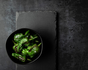 An overhead view of a small bowl of blistered padron peppers with maldon sea salt shot on a dark surface