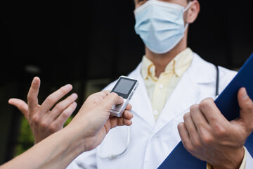 cropped view of reporter holding voice recorder near doctor in medical mask gesturing during...