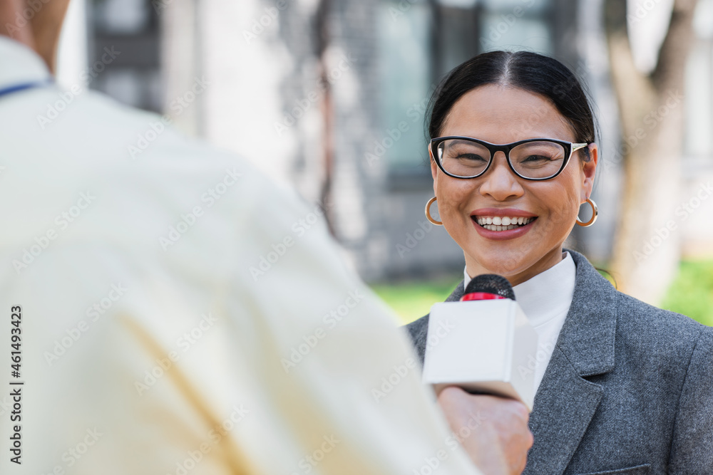 Poster blurred reporter standing with microphone near positive asian businesswoman in glasses