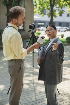 journalist holding microphone and taking interview of asian businesswoman in glasses holding clipboard and pointing away
