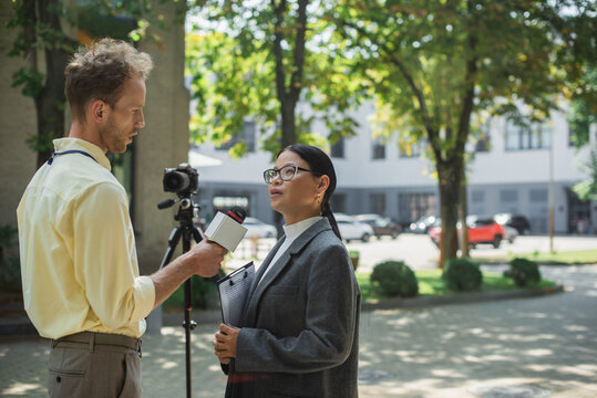 journalist holding microphone and taking interview of asian businesswoman in glasses near digital camera on street