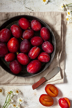 A vintgae tin filled with homegrown red plums resting on a folden cloth with a cut plum beside it.