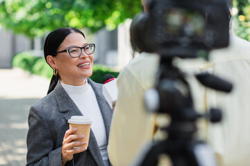 blurred reporter holding microphone near happy asian businesswoman with paper cup giving interview - obrazy, fototapety, plakaty