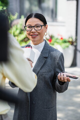 cheerful asian businesswoman in glasses holding smartphone and giving interview near blurred...