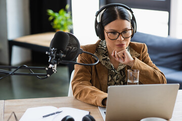 pensive asian radio host in eyeglasses and headphones using laptop near near microphone with pop...
