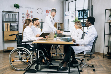 Obraz premium Bearded caucasian doctor shaking hands and greeting his multi ethnic colleagues during medical seminar at office. Female wheelchair user working with colleagues on common project.